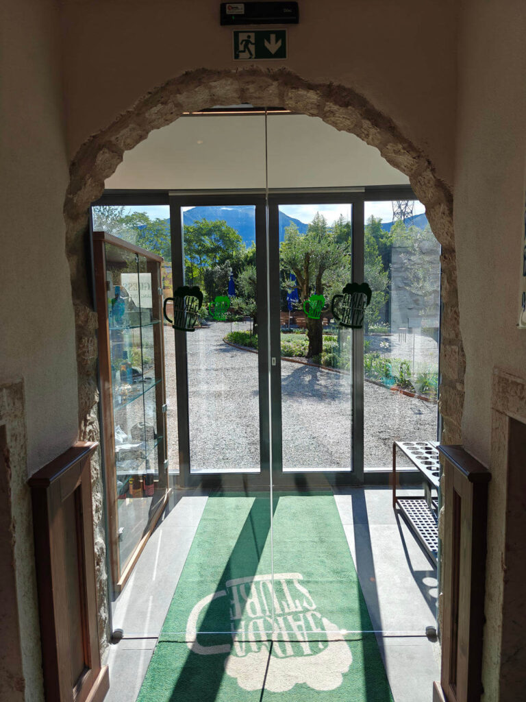 Design Luminoso e Funzionale: I Nostri Serramenti per Garda Stube di Riva del Garda (TN) Immagine interna di un ingresso moderno in una struttura ricettiva, il ristorante Garda Stube a Riva del Garda (TN), con un'ampia porta a vetri che si apre su un'area esterna. La porta è incorniciata da un arco in pietra rustica, e sul pavimento si trova uno zerbino verde con il logo "GARDA STUBE". Attraverso il vetro, si vedono un vialetto di ghiaia, alberi di ulivo e la vegetazione circostante con montagne sullo sfondo. Un carrello nero è visibile sulla destra, e una vetrina sulla sinistra.