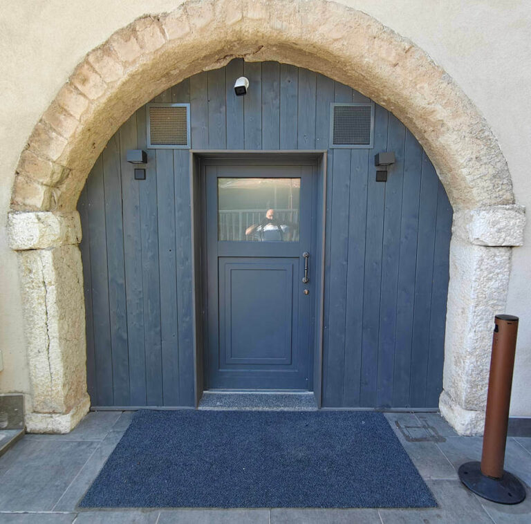 Portone in Legno e Vetro per Ristorante a Riva del Garda: Garda Stube (TN) Immagine ravvicinata di un ingresso in legno scuro, incorniciato da un grande arco in pietra rustica e situato in un edificio a Riva del Garda (TN), questo è il ristorante Garda Stube. La porta principale è di colore grigio scuro, con un pannello di vetro superiore, e presenta una maniglia in metallo cromato. Sopra la porta, sono visibili due griglie di ventilazione e un riflettore scuro. A pavimento, davanti all'ingresso, si trova uno zerbino di colore blu scuro.