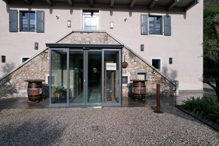Chiusure e Ingressi in Vetro per Ristoranti e Alberghi a Riva del Garda, Trentino: Garda Stube Immagine esterna di una sezione di un edificio a Riva del Garda (TN), il Garda Stube ristorante tipico Bavarese e birrificio con una facciata chiara e finestre con scuri grigi a battente. La parte centrale dell'edificio presenta un ingresso moderno in vetro e infissi grigi, incassato in una base di pietra. Due scalinate laterali in pietra conducono all'ingresso superiore. Davanti all'ingresso, sono posizionate due botti di legno che fungono da tavolini, e il pavimento è in ghiaia chiara. Sullo sfondo, si intravedono alberi e la vegetazione tipica della zona.