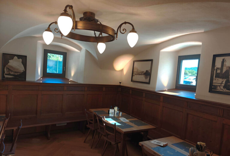 Realizzazione Serramenti Garda Stube Riva del Garda (TN): Interni Accoglienti e Illuminazione Ambiente Immagine interna di un ambiente di ristorante, il Garda Stube a Riva del Garda (TN), caratterizzato da boiserie in legno scuro che riveste la parte inferiore delle pareti. Le pareti superiori sono di colore bianco e presentano nicchie arcuate che contengono finestre rettangolari. Due finestre mostrano la vista del verde esterno e una terza finestra è parzialmente visibile a destra. Il soffitto è bianco, con un grande lampadario circolare in ferro battuto con cinque lampade a forma di goccia che pende al centro. Tavoli in legno con panche e sedie sono disposti nell'ambiente, e alcuni quadri sono appesi alle pareti.