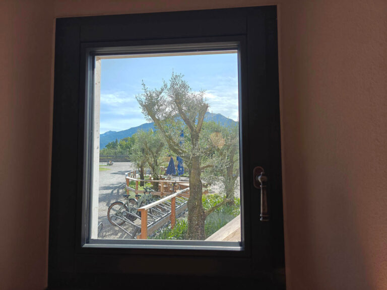 Infissi e Chiusure per Ristoranti a Riva del Garda, Trentino: Ristorante Garda Stube (TN) Vista attraverso una finestra quadrata con infissi scuri, interno del ristorante Garda Stube a Riva del Garda (TN) che mostra un'area esterna con alberi di ulivo, tavoli e sedie. Sullo sfondo, si distinguono montagne verdi sotto un cielo azzurro e parzialmente nuvoloso. All'interno, parte della parete di colore rosa chiaro e una maniglia in metallo scuro sul lato destro della finestra sono visibili. Un ombrellone blu è parzialmente visibile dietro gli alberi.