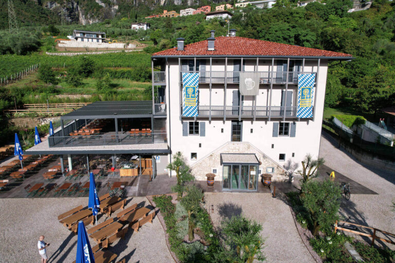 Realizzazione Serramenti Garda Stube Riva del Garda (TN): Architettura Tradizionale e Design Funzionale Veduta aerea di un edificio a più piani, identificato come "GardaStube" a Riva del Garda (TN), con tetto in tegole rosse e facciate chiare. L'edificio presenta ampi balconi in legno con parapetti a ringhiera. Sulle facciate sono appese delle bandiere a scacchi blu e bianchi. In primo piano, c'è un'ampia area esterna con tavoli e panche in legno, ombrelloni blu aperti e delle piante. Sulla sinistra, si trova un'altra struttura con tetto scuro e area esterna attrezzata per la ristorazione. Sullo sfondo, si estendono colline verdi e un vigneto.