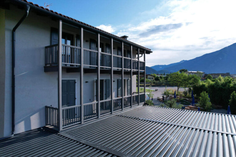 Realizzazione Serramenti GardaS tube Riva del Garda (TN): Terrazza Panoramica e Pergola Bioclimatica Immagine esterna di una sezione di un edificio moderno, il Garda Stube a Riva del Garda (TN), ristorante tipico con specialità bavaresi e birra. E' stata ripresa con il drone una lunga terrazza coperta da una pergola a lamelle orizzontali di colore scuro. Il primo piano dell'edificio presenta balconi con ringhiere grigie e finestre con scuri grigi. Sotto la pergola, si intravede un'area esterna con tavoli e ombrelloni blu. Il tetto dell'edificio è in tegole rosse. Sullo sfondo, si stagliano imponenti montagne sotto un cielo azzurro con nuvole sparse.