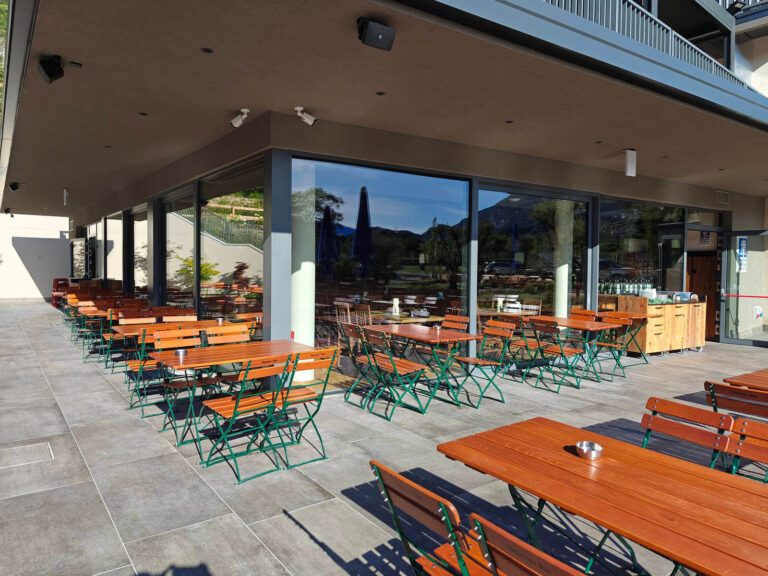 Design Luminoso e Funzionale: I Nostri Serramenti per la Garda Stube di Riva del Garda (TN) Immagine esterna di un moderno ristorante, il Garda Stube a Riva del Garda (TN), con una lunga facciata in vetro che riflette l'ambiente circostante. Numerosi tavoli e sedie in legno con telai verdi sono disposti su un'ampia terrazza pavimentata, pronti per gli ospiti. In lontananza, attraverso le vetrate, si intravedono alberi e montagne. Il soffitto della terrazza è di colore scuro, con faretti incassati e telecamere di sorveglianza visibili.