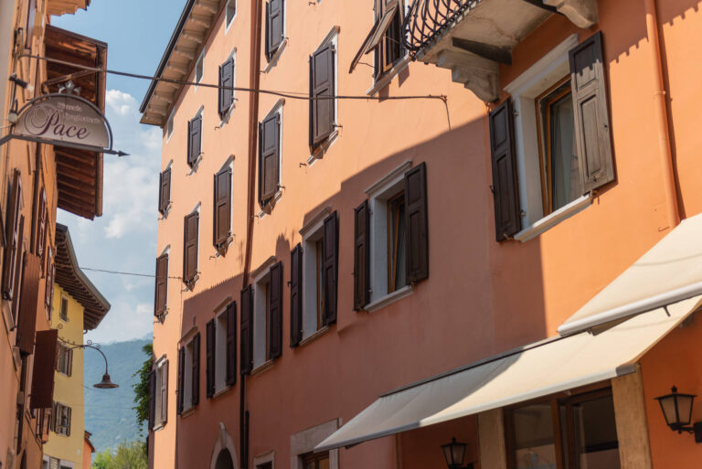 Design Tradizionale e Infissi di Qualità: I Nostri Serramenti per l’Hotel Pace di Arco (TN) Veduta esterna di un edificio a più piani con facciata arancione chiaro, è l'Hotel Pace nel centro storico di Arco (TN). L'edificio presenta numerose finestre con persiane in legno scuro e un balcone in ferro battuto decorato. Sulla sinistra, è visibile un'insegna circolare appesa con la scritta "Pace". In primo piano a destra, si nota una tenda da sole chiara sopra un'altra finestra. Sullo sfondo si intravedono altri edifici e colline.