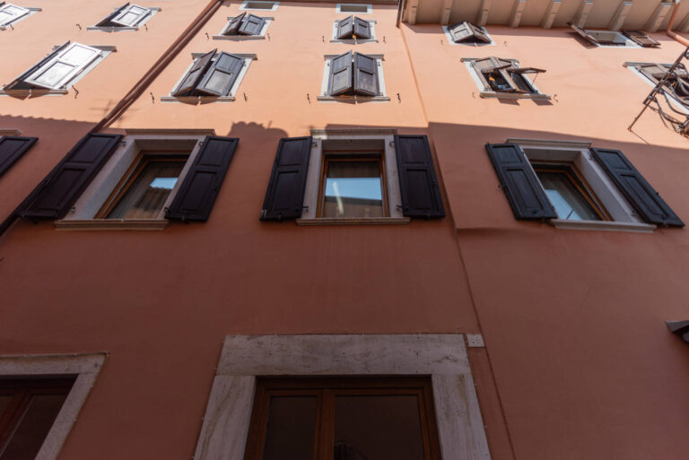 Realizzazione Serramenti Hotel Pace Arco (TN): Facciata Storica e Finestre Moderne nel Cuore del Centro Immagine ravvicinata della facciata esterna di un edificio, l'Hotel Pace nel centro storico di Arco (TN). La facciata è di colore arancione chiaro e presenta numerose finestre con persiane in legno scuro, alcune delle quali sono aperte a varie angolazioni. Le finestre hanno cornici chiare e riflettono il cielo. La parte inferiore della facciata mostra una base in pietra chiara e un portone in legno.