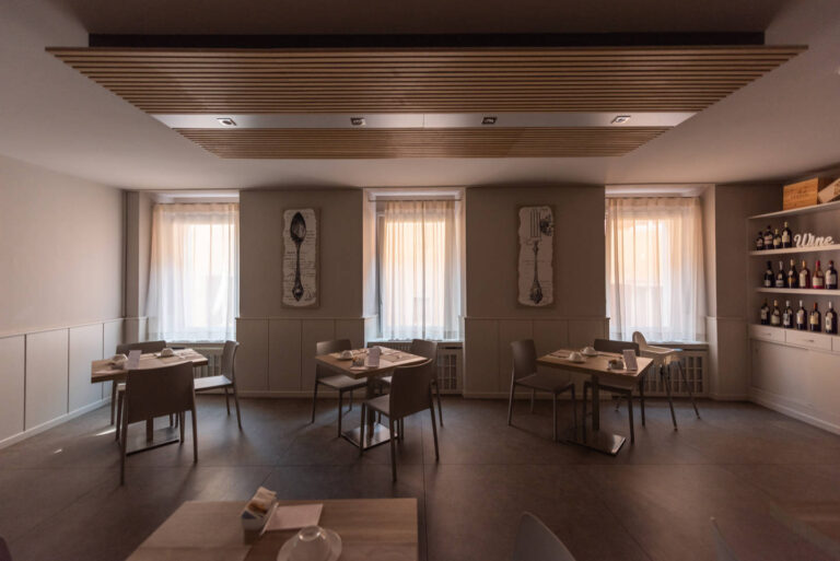 Realizzazione Serramenti Hotel Pace Arco (TN): Sala Colazioni Moderna con Ambienti Luminosi Immagine interna di una moderna sala colazioni all'Hotel Pace ad Arco (TN). L'ambiente è arredato con tavoli quadrati e sedie grigie, disposti ordinatamente. Le pareti sono chiare, con una boiserie orizzontale nella parte inferiore e tre finestre rettangolari con tende bianche trasparenti che lasciano filtrare la luce naturale. Il soffitto presenta un design a doghe di legno chiare con illuminazione integrata, e una mensola sulla destra mostra bottiglie di vino.