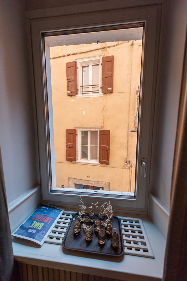 Design Funzionale e Isolamento Acustico: I Nostri Serramenti per l’Hotel Pace di Arco (TN) Vista dall'interno di una camera d'albergo, presso Hotel Pace nel centro storico di Arco (TN), vista attraverso una finestra rettangolare con infissi bianchi, che affaccia su un edificio di fronte. L'edificio esterno ha una facciata gialla con due finestre visibili, entrambe con scuri marroni aperti. Sul davanzale della finestra interna sono posizionati una brochure a tema "BIKE" e un vassoio scuro con piccole decorazioni floreali secche. Delle tende marroni sono visibili ai lati della finestra.
