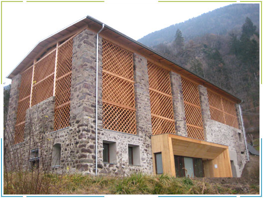 Design Rurale Contemporaneo: Le Nostre Chiusure per il Maso Hall di Merano (Alto Adige) Veduta esterna di un edificio rustico, il Maso Hall a Merano (BZ), con facciata in pietra e ampie sezioni di rivestimento in legno a griglia. L'edificio presenta un tetto spiovente e una struttura lignea sotto la gronda. Al piano terra, un moderno ingresso in legno chiaro sporge dalla facciata in pietra, con finestre scure sottostanti. Il terreno circostante è leggermente in pendenza con vegetazione. Sullo sfondo si ergono montagne verdi e un cielo nuvoloso.