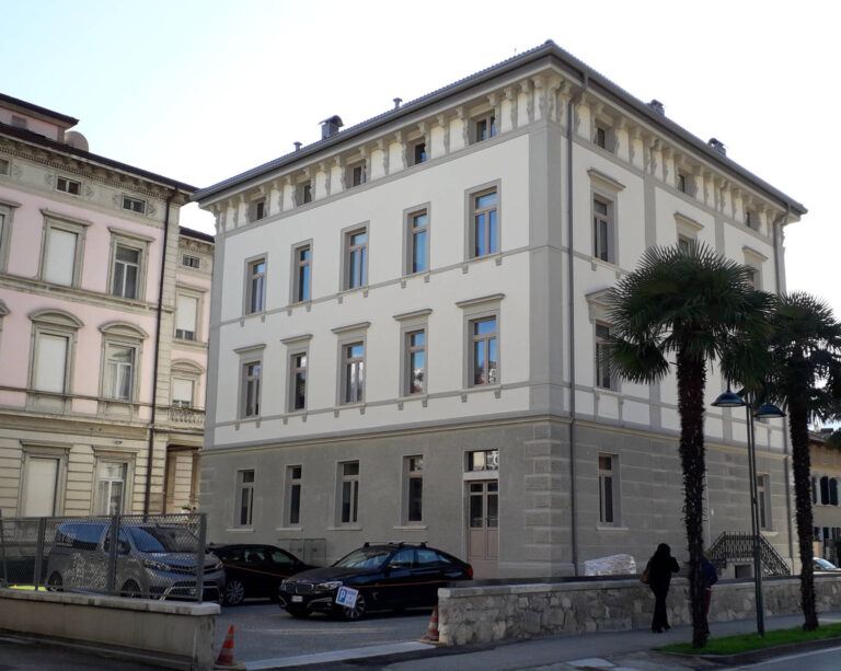 Serramenti di Alta Qualità per Edifici Storici: Residenza Alto Garda, Riva del Garda (TN) Veduta esterna di un imponente edificio storico a più piani con facciata bianca e elementi decorativi grigi, la Residenza Alto Garda a Riva del Garda (TN). L'edificio è ad angolo e presenta finestre con cornici e balconi in ferro battuto. Sul lato destro, due alte palme aggiungono un tocco mediterraneo all'ambiente. Accanto ad esso, sulla sinistra, si intravede un altro edificio simile di colore rosa chiaro, probabilmente il prestigioso Hotel Liberty. In primo piano, c'è una strada con alcune auto parcheggiate e un muretto in pietra.