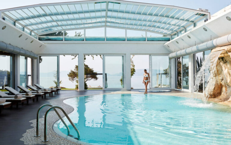 Piscina Resort Lusso con Vista Mare: Serramenti su Misura per Hydra Beach SA, Mar Egeo Immagine interna di una grande piscina coperta in un resort di lusso, l'Hydra Beach SA in Grecia. La piscina, di forma irregolare, è circondata da pavimenti scuri e lettini prendisole. Un'ampia vetrata con infissi bianchi si estende lungo tutta la parete, offrendo una vista panoramica sul mare e sulla vegetazione esterna. Il soffitto è caratterizzato da una struttura vetrata inclinata che lascia filtrare abbondante luce naturale. Sulla destra, una cascata d'acqua scende da una formazione rocciosa artificiale.