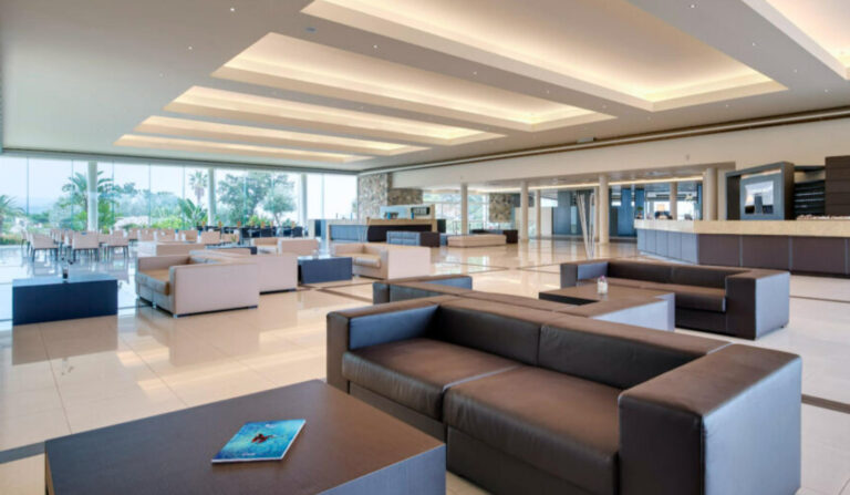 Interni di Lusso e Spazi Luminosi per Hotel in Grecia: Hydra Beach SA (Mar Egeo) Immagine interna di un'ampia e moderna lobby di un resort, l'Hydra Beach SA in Grecia. Lo spazio è caratterizzato da pavimenti lucidi di colore chiaro e un soffitto con illuminazione a incasso. Numerosi divani in pelle scura e poltrone chiare sono disposti con tavolini bassi. Una grande vetrata si estende lungo la parete di fondo, offrendo una vista su una zona esterna con palme e altre sedute. Sul lato destro, si intravede un'area reception con bancone.