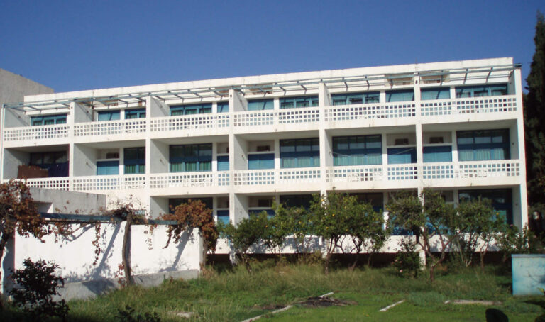 Design Funzionale e Luminoso: I Nostri Serramenti per il Resort Thermisia (Grecia) Veduta esterna di un moderno edificio bianco a più piani, il Resort Thermisia in Grecia. La facciata è caratterizzata da una serie di balconi aperti con ringhiere bianche e finestre con infissi turchesi. Sopra ogni fila di balconi, una struttura a griglia in metallo chiaro fornisce ombra. In primo piano, c'è un'area verde con alberi e arbusti.