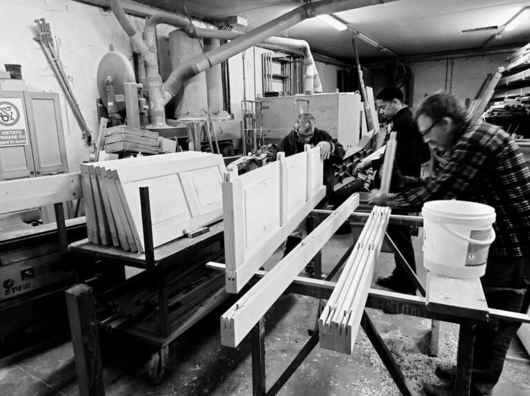 Immagine in bianco e nero che cattura l'attività all'interno del nostro laboratorio di falegnameria ProBauen, dove tre uomini adulti sono intenti a lavorare su elementi di porte o mobili in legno. In primo piano a sinistra, si vedono diverse sezioni di pannelli di legno impilati, alcuni dei quali sono di colore chiaro, probabilmente già trattati. Al centro, un uomo in primo piano sta piegato su un pezzo di legno, con le mani impegnate in un'azione specifica, mentre un altro uomo in piedi sulla destra, con una camicia a quadri, sta lavorando su un altro elemento di legno vicino a un secchio bianco. Un terzo uomo è visibile più in profondità nel laboratorio, anch'esso impegnato. L'ambiente è tipico di una falegnameria, con macchinari industriali, tubi di aspirazione e attrezzi sparsi, suggerendo un'attività artigianale e industriale.