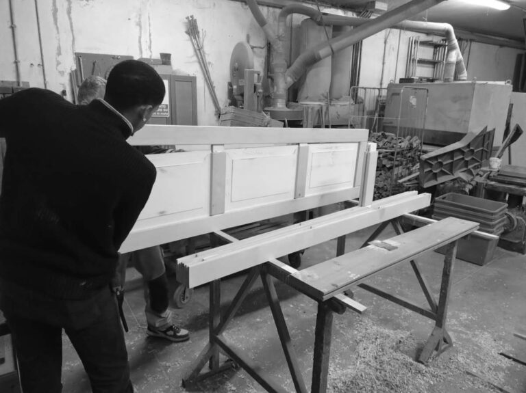 Immagine in bianco e nero che riprende da dietro un falegname con un maglione scuro, chino su un pezzo di legno chiaro appoggiato su cavalletti. Sta lavorando a un pannello o una sezione di porta che presenta tre riquadri. Sulla destra del pannello in lavorazione si trovano altri elementi di legno più lunghi, allineati sui cavalletti. L'ambiente è un laboratorio di falegnameria, visibilmente attrezzato con macchinari industriali, tubi di aspirazione e vari attrezzi e materiali sparsi, suggerendo un contesto di lavoro artigianale e industriale. Sul pavimento, si notano trucioli e segatura. Un'altra persona è parzialmente visibile in sottofondo, vicino ai macchinari.