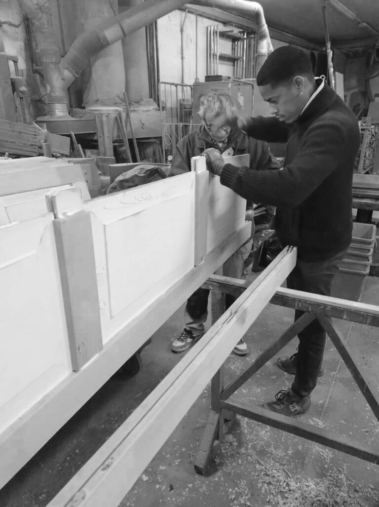 Immagine in bianco e nero che mostra due falegnami impegnati nella fase di assemblaggio di un elemento in legno, probabilmente parte di una porta o di un mobile. Entrambi gli artigiani sono chinati sul pezzo di legno, che appare di colore chiaro e ben rifinito, appoggiato su cavalletti. La persona in primo piano a destra indossa un maglione scuro e guanti, ed è concentrata sul lavoro manuale. Sullo sfondo si intravedono attrezzature e macchinari tipici di un laboratorio di falegnameria, con trucioli di legno sparsi sul pavimento, che indicano un ambiente di lavoro attivo e artigianale.