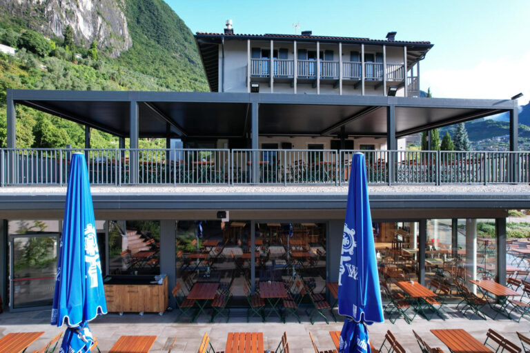 Design Luminoso e Spazi Funzionali: I Nostri Serramenti per il Garda Stube di Riva del Garda (TN) Immagine esterna di un moderno ristorante, il Garda Stube a Riva del Garda (TN), con una grande vetrata al piano terra che mostra l'interno con tavoli e sedie in legno. All'esterno, in primo piano, si estende un'ampia area pavimentata con numerosi tavoli e sedie in legno, alcuni con ombrelloni blu aperti. Al piano superiore, una grande terrazza con ringhiere metalliche grigie è visibile, parzialmente coperta da una struttura scura con lamelle. Sullo sfondo, si ergono montagne verdi sotto un cielo azzurro e nuvoloso.