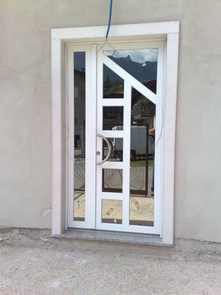 Porta d’Ingresso Design in Vetro a Drena (TN): Soluzioni Serramenti Alta Qualità Immagine verticale di una porta d'ingresso moderna con ampie superfici vetrate, incorniciata in una parete di colore chiaro. La porta è di colore bianco o grigio chiaro, con un'anta principale e un pannello laterale stretto, entrambi dotati di vetri che riflettono il paesaggio circostante, inclusi edifici e montagne. L'anta principale presenta un design geometrico con barre diagonali e orizzontali e una maniglia a mezzaluna in metallo lucido. La porta è incorniciata da uno stipite esterno in pietra chiara. Sullo sfondo, attraverso il vetro, si intravedono elementi urbani e naturali del luogo, siamo a Drena (TN).