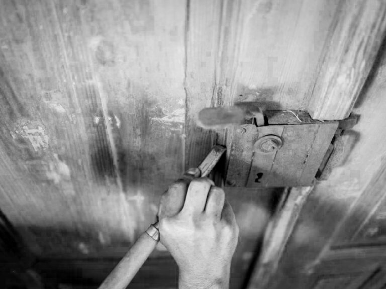 Questa immagine in bianco e nero mostra una persona che lavora su una vecchia porta di legno grezza. Si intravede la parte superiore del corpo e le mani della persona che sono impegnate con un attrezzo, forse per rimuovere o riparare una serratura metallica arrugginita che si trova sul lato destro dell'immagine. La grana del legno della porta è molto evidente, con linee verticali e imperfezioni naturali. L'illuminazione crea contrasti e ombre, enfatizzando la texture sia del legno che del metallo.
