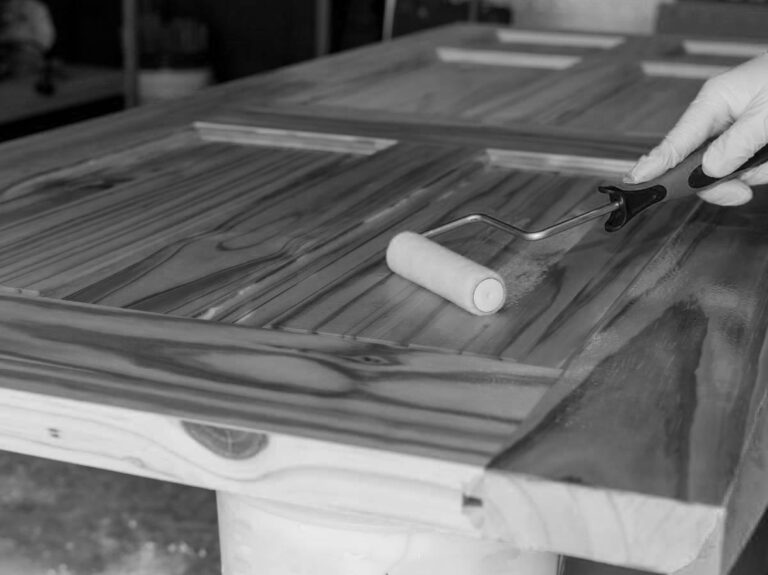 Immagine in bianco e nero che mostra una persona con un guanto bianco che applica una finitura trasparente su una porta di legno restaurata, appoggiata orizzontalmente. La persona tiene un piccolo rullo per vernice, che sta passando su una delle sezioni a pannelli della porta. Il legno sottostante ha una venatura molto evidente e scura, e la parte già trattata appare più lucida rispetto al legno non ancora finito. L'immagine si concentra sulla porta e sull'azione di finitura.