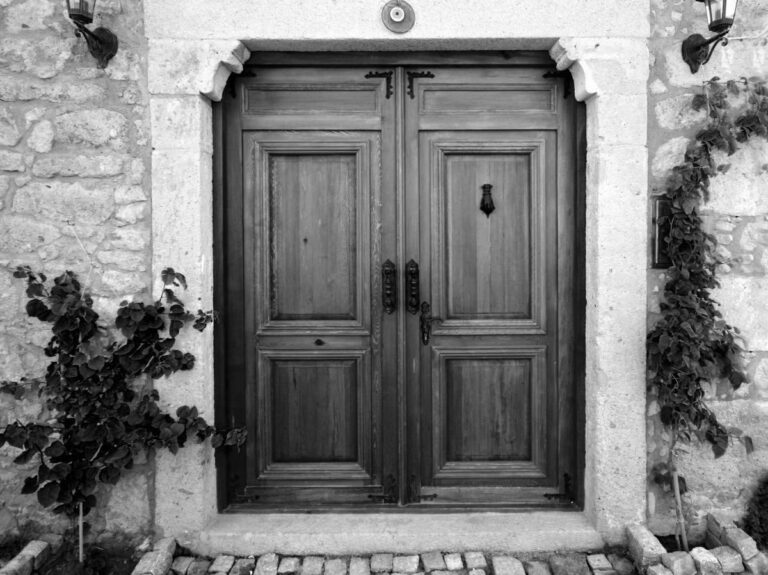 Immagine in bianco e nero di un imponente portone antico in legno a due ante, restaurato, incastonato in una facciata di mattoni o pietre grezze. Il portone presenta pannelli rettangolari in rilievo su ciascuna anta e dettagli decorativi in ferro battuto, inclusi cardini ornamentali nella parte superiore. Sull'anta destra si nota un battente a forma di anello scuro. Il portone è incorniciato da uno stipite in pietra lavorata con dettagli curvi sull'architrave. Ai lati del portone, sulle pareti in pietra, sono appese due lanterne in ferro battuto. Delle piante rampicanti e arbusti crescono ai lati dell'ingresso. Il portone poggia su un gradino di pietra e un selciato irregolare.