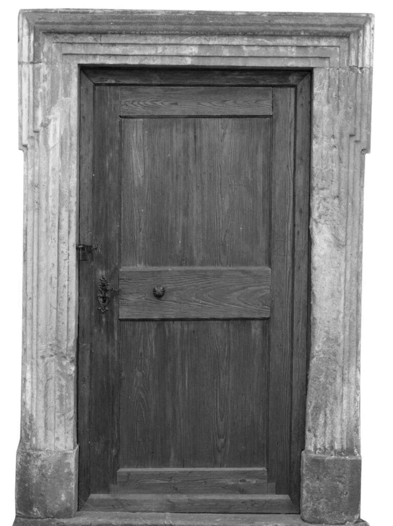 Immagine in bianco e nero di una porta in legno antica, restaurata, incorniciata da un imponente stipite in pietra scolpita. La porta è composta da pannelli di legno verticale con una venatura ben visibile, e presenta un pannello orizzontale all'altezza della maniglia, che ha un pomello rotondo e una piastra decorata. Sul lato sinistro della porta è visibile una serratura o un chiavistello in ferro battuto. Lo stipite in pietra ha una lavorazione a colonne scanalate e un architrave anch'esso decorato, che conferisce alla porta un aspetto nobile e antico. Lo sfondo è uniformemente bianco, isolando la porta e il suo contesto immediato.