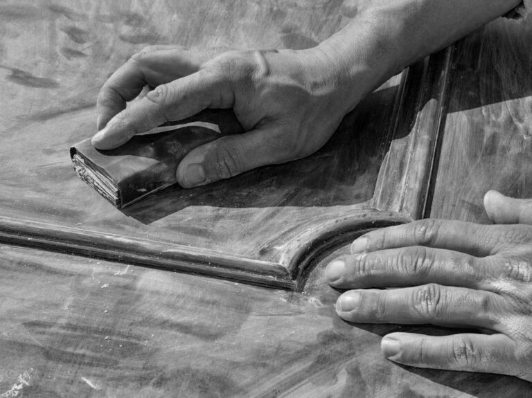 L’Arte del Restauro: Levigatura di Dettagli Intagliati Immagine in bianco e nero che mostra da vicino le mani di una persona mentre levigano la superficie di un vecchio portone o pannello in legno. La mano sinistra tiene un blocco di carta vetrata piegata, che sta passando con cura su un bordo intagliato del legno. La mano destra è appoggiata sulla superficie del legno per stabilizzare. Il legno appare grezzo e mostra segni di polvere di levigatura, indicando che il lavoro è in corso. La venatura del legno è visibile e conferisce profondità alla texture.