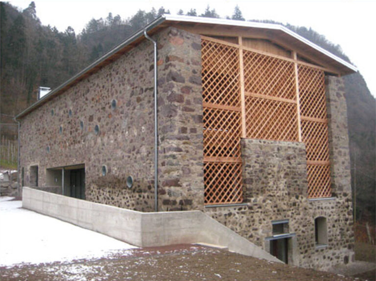 Innovazione e Tradizione: Serramenti e Chiusure di Design per Architetture Uniche a Merano (BZ) Edificio dal design distintivo con pareti in pietra e ampie sezioni rivestite da innovative chiusure a griglia in legno chiaro. In basso, si nota un moderno ingresso con vetrate e una pensilina in legno. L'ambiente è montano, suggerendo l'applicazione di serramenti di alta qualità per combinare estetica e performance in contesti unici.